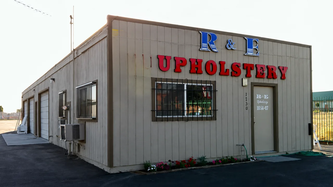 The R&E Upholstery shop in Bloomington, California: a single-storey building with the red and blue R&E Upholstery sign, three roll-up bay doors and flowers along the entrance